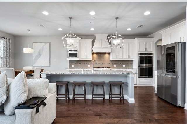 kitchen with appliances with stainless steel finishes, custom range hood, a kitchen island with sink, dark wood-type flooring, and pendant lighting