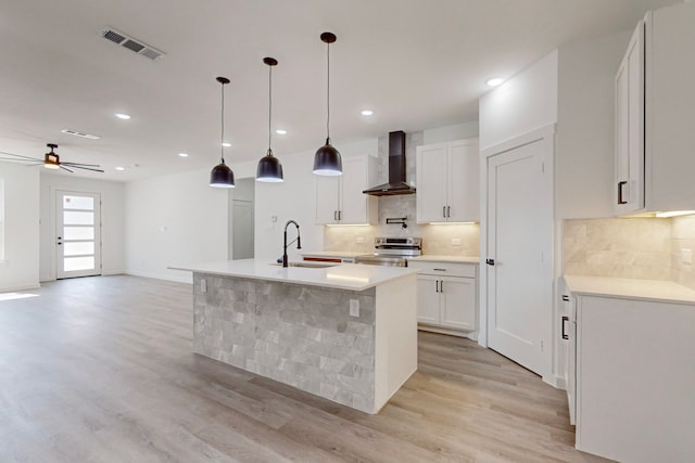 kitchen featuring wall chimney exhaust hood, decorative light fixtures, stainless steel electric range oven, a kitchen island with sink, and light wood-type flooring