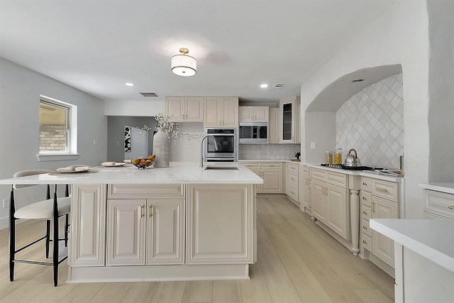 kitchen featuring a kitchen island with sink, backsplash, appliances with stainless steel finishes, light wood-type flooring, and a kitchen bar