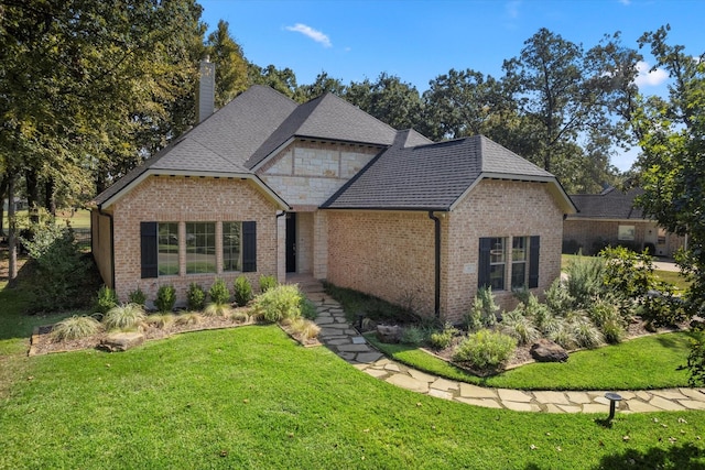 view of front facade featuring brick siding, roof with shingles, a chimney, a front yard, and stone siding