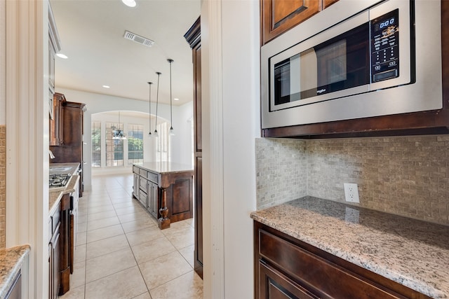 kitchen featuring light stone counters, tasteful backsplash, stainless steel microwave, light tile patterned flooring, and decorative light fixtures