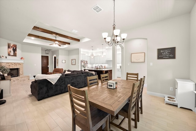 dining space featuring a tray ceiling, beamed ceiling, ceiling fan with notable chandelier, a fireplace, and light hardwood / wood-style floors