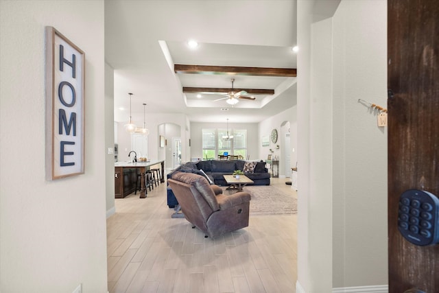 living room featuring sink, light wood-type flooring, ceiling fan with notable chandelier, a raised ceiling, and beam ceiling