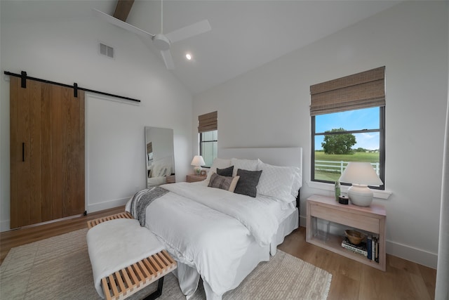 bedroom with a barn door, ceiling fan, light hardwood / wood-style flooring, and high vaulted ceiling