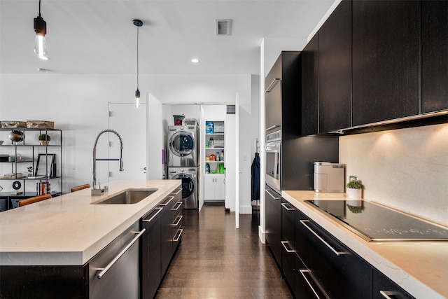 kitchen featuring dark wood-type flooring, appliances with stainless steel finishes, stacked washer / drying machine, decorative light fixtures, and a center island with sink