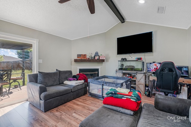living room featuring a ceiling fan, a textured ceiling, wood finished floors, a glass covered fireplace, and vaulted ceiling with beams