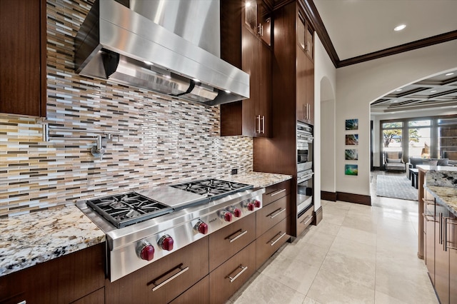 kitchen featuring stainless steel appliances, light stone countertops, ornamental molding, wall chimney range hood, and decorative backsplash