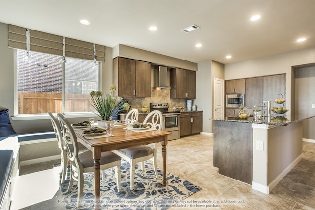 kitchen featuring dark brown cabinetry, dark stone countertops, wall chimney range hood, appliances with stainless steel finishes, and backsplash