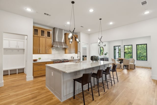 kitchen featuring a large island with sink, pendant lighting, light stone countertops, wall chimney exhaust hood, and light hardwood / wood-style flooring