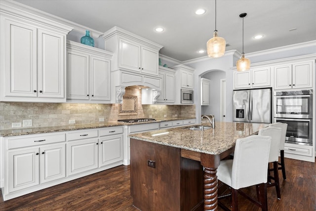 kitchen with ornamental molding, stainless steel appliances, a kitchen island with sink, white cabinets, and hanging light fixtures