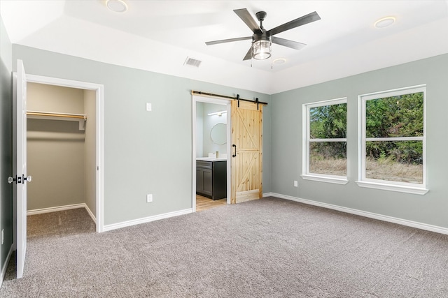 unfurnished bedroom featuring a barn door, ceiling fan, light carpet, a spacious closet, and ensuite bath