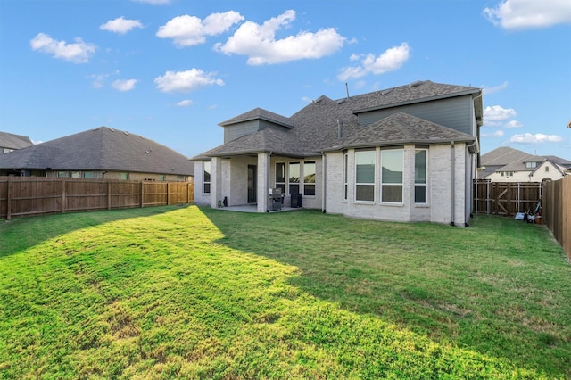 back of house with a shingled roof, a patio, a fenced backyard, a yard, and brick siding