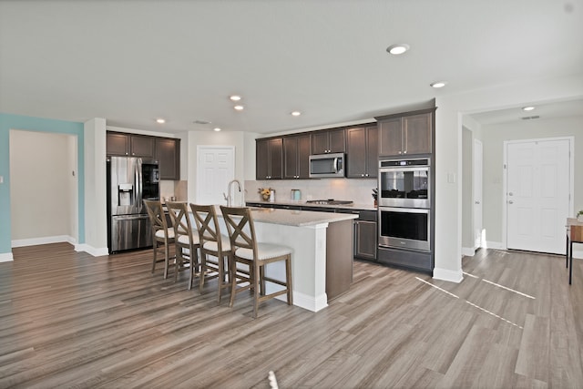 kitchen featuring appliances with stainless steel finishes, backsplash, a breakfast bar, a center island with sink, and light hardwood / wood-style flooring