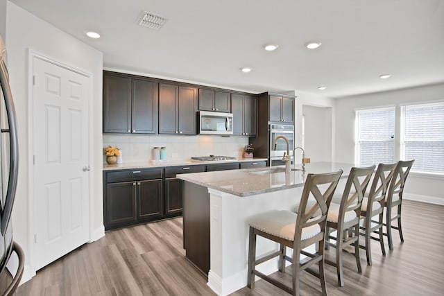 kitchen featuring sink, stainless steel appliances, light hardwood / wood-style flooring, backsplash, and a center island with sink