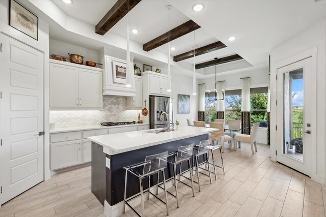 kitchen with appliances with stainless steel finishes, beamed ceiling, a kitchen island with sink, and white cabinetry