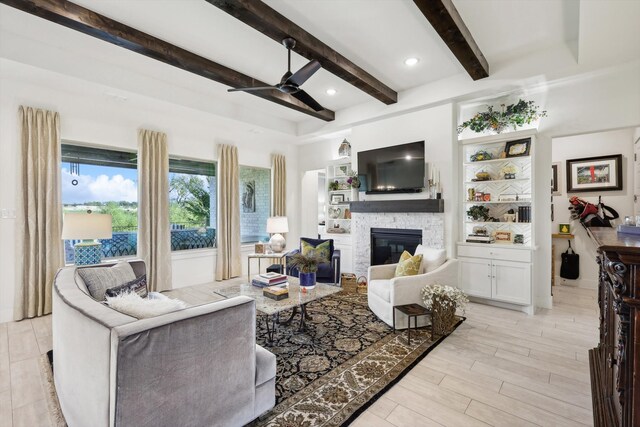 living room featuring ceiling fan, beamed ceiling, light hardwood / wood-style flooring, and a stone fireplace