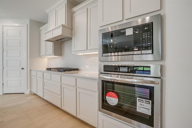 kitchen with light wood-type flooring, tasteful backsplash, custom range hood, white cabinets, and appliances with stainless steel finishes