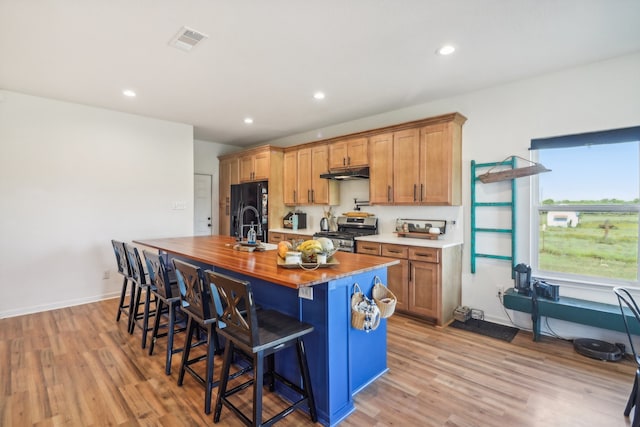 kitchen with a kitchen bar, light wood-type flooring, black refrigerator with ice dispenser, a center island with sink, and stainless steel range oven