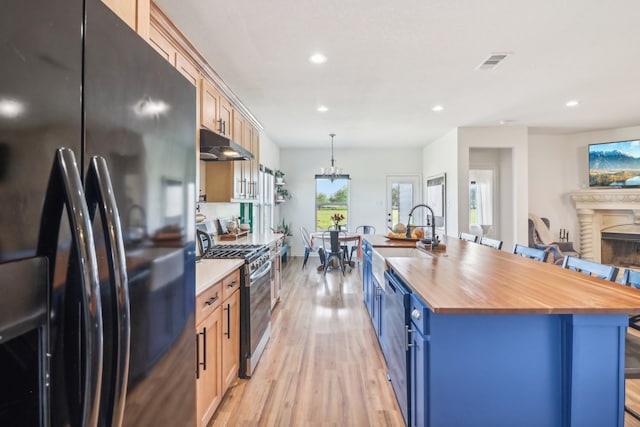 kitchen with wooden counters, light wood-type flooring, a kitchen island with sink, black appliances, and hanging light fixtures