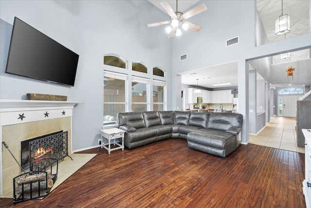 living room featuring ceiling fan, a tile fireplace, a towering ceiling, and light hardwood / wood-style flooring