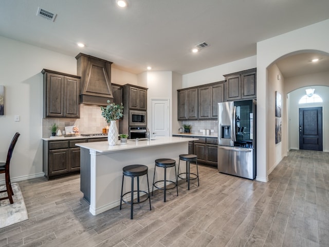 kitchen with custom exhaust hood, light wood-type flooring, stainless steel appliances, and a kitchen island with sink