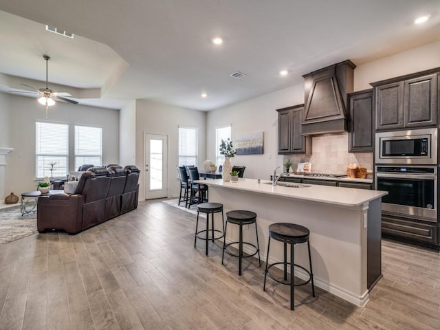 kitchen with light wood-type flooring, custom range hood, stainless steel appliances, sink, and a center island with sink