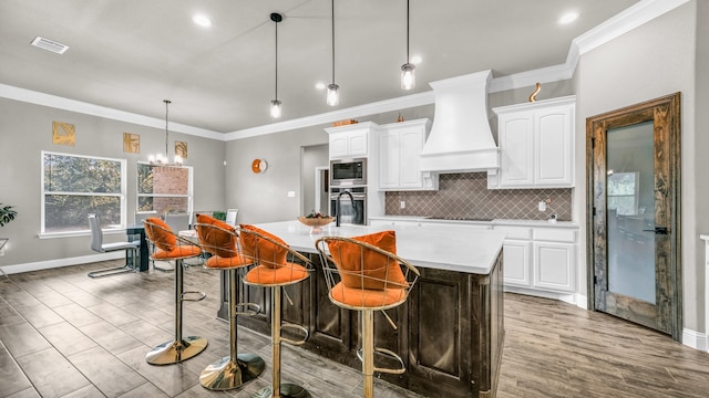 kitchen featuring an island with sink, custom range hood, pendant lighting, white cabinetry, and appliances with stainless steel finishes