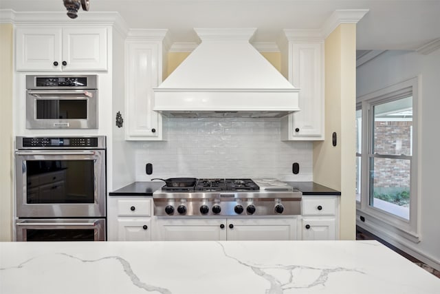 kitchen with tasteful backsplash, dark stone countertops, custom exhaust hood, white cabinets, and appliances with stainless steel finishes