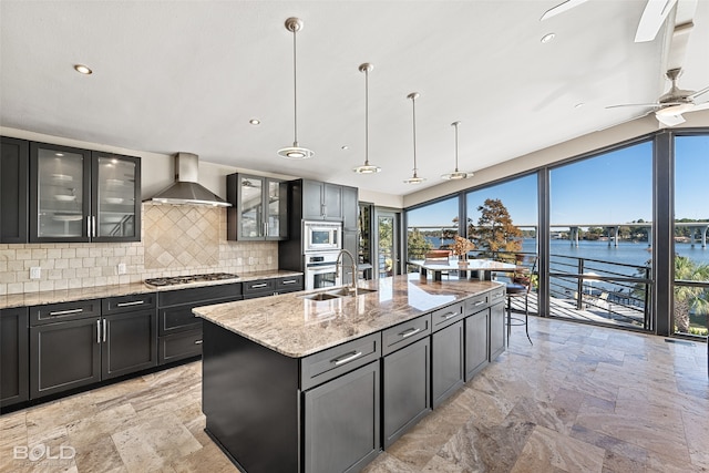 kitchen with decorative backsplash, wall chimney exhaust hood, hanging light fixtures, appliances with stainless steel finishes, and light stone counters