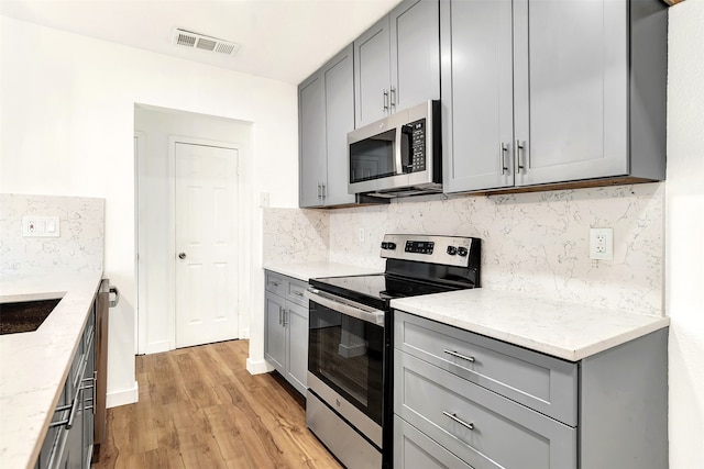 kitchen featuring gray cabinetry, light stone countertops, light wood-type flooring, stainless steel appliances, and decorative backsplash