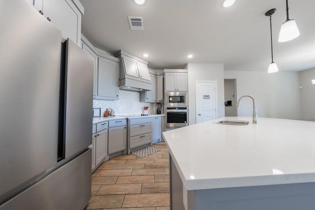 kitchen featuring a kitchen island with sink, custom range hood, sink, hanging light fixtures, and appliances with stainless steel finishes