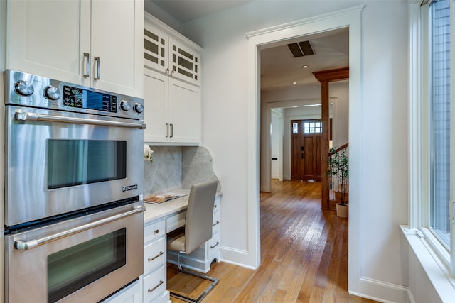 kitchen featuring built in desk, white cabinetry, stainless steel double oven, light hardwood / wood-style floors, and decorative backsplash