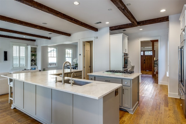 kitchen with beamed ceiling, light wood-type flooring, sink, an island with sink, and appliances with stainless steel finishes