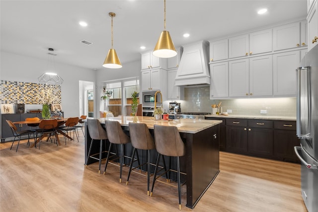 kitchen with a center island with sink, custom exhaust hood, pendant lighting, and white cabinetry