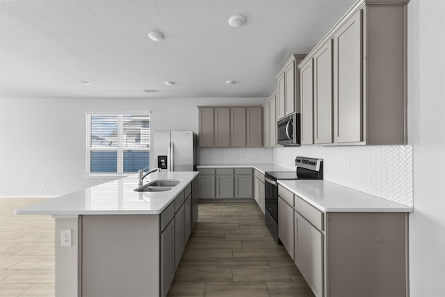 kitchen with an island with sink, sink, backsplash, gray cabinets, and stainless steel appliances