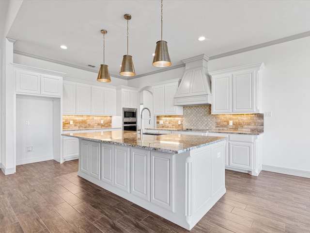 kitchen with premium range hood, white cabinetry, appliances with stainless steel finishes, and an island with sink