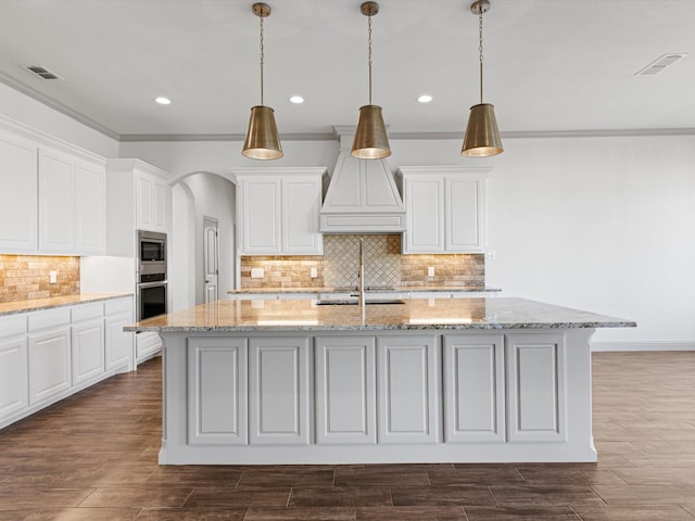 kitchen featuring premium range hood, appliances with stainless steel finishes, decorative light fixtures, and an island with sink