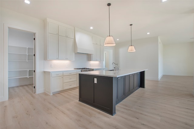 kitchen with premium range hood, an island with sink, decorative light fixtures, white cabinetry, and light wood-type flooring