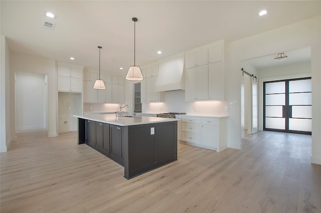 kitchen with a barn door, light hardwood / wood-style floors, custom range hood, an island with sink, and white cabinets