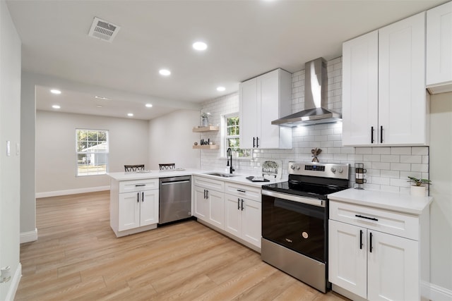 kitchen with light wood-type flooring, stainless steel appliances, sink, wall chimney range hood, and white cabinetry