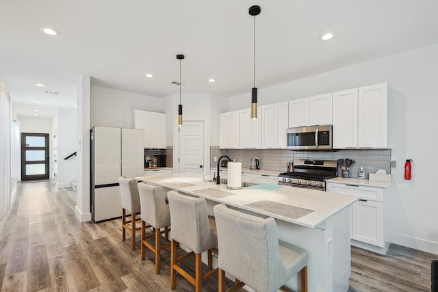 kitchen featuring an island with sink, stainless steel appliances, pendant lighting, white cabinetry, and light hardwood / wood-style floors