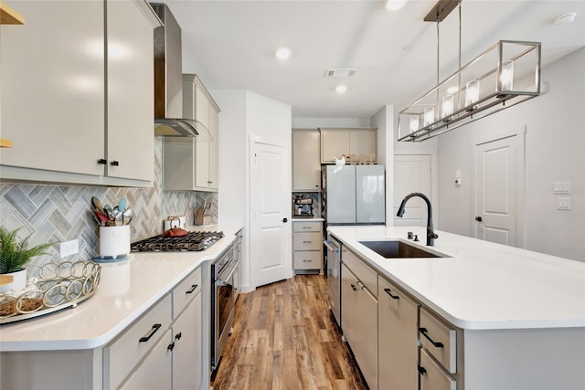 kitchen with hardwood / wood-style flooring, wall chimney exhaust hood, a center island with sink, sink, and appliances with stainless steel finishes