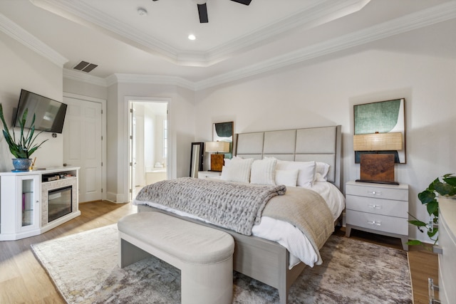 bedroom with light wood-type flooring, ensuite bath, a raised ceiling, ceiling fan, and ornamental molding