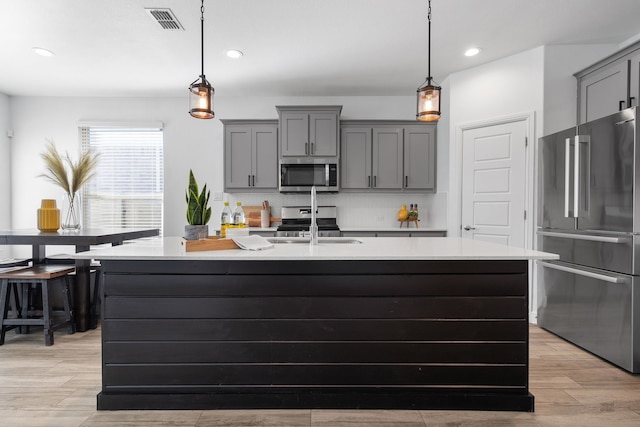 kitchen with decorative backsplash, an island with sink, stainless steel appliances, gray cabinetry, and decorative light fixtures