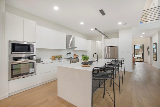 kitchen featuring wall chimney range hood, stainless steel appliances, decorative light fixtures, white cabinets, and a kitchen island with sink