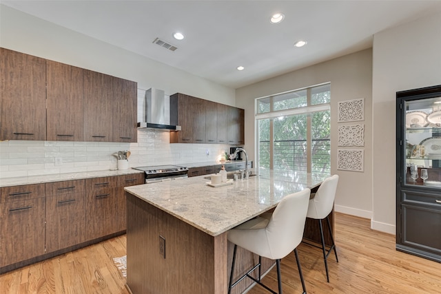 kitchen with sink, light wood-type flooring, an island with sink, stainless steel range with electric stovetop, and wall chimney exhaust hood