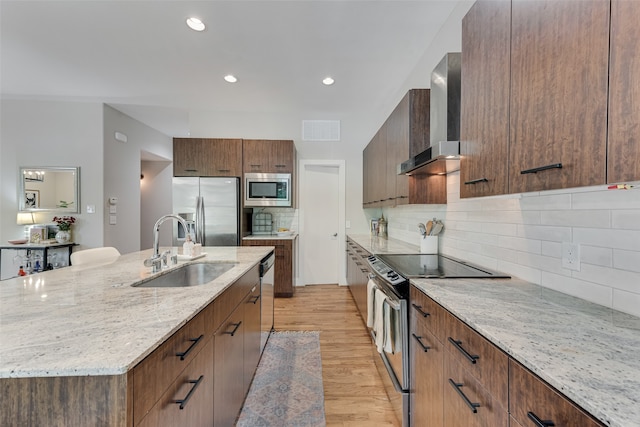 kitchen featuring wall chimney range hood, sink, an island with sink, light hardwood / wood-style floors, and stainless steel appliances