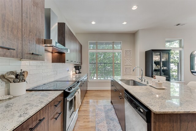 kitchen with light wood-type flooring, stainless steel appliances, wall chimney exhaust hood, light stone counters, and a center island with sink