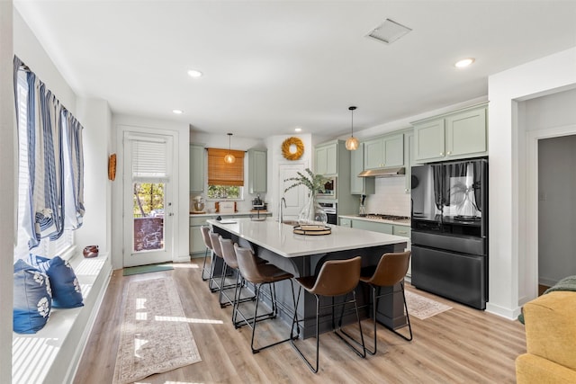 kitchen featuring light wood-type flooring, backsplash, stainless steel appliances, a kitchen island with sink, and hanging light fixtures