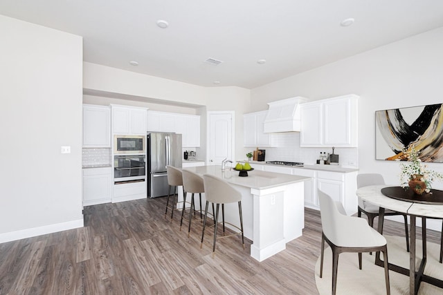 kitchen featuring appliances with stainless steel finishes, white cabinetry, decorative backsplash, a kitchen island with sink, and custom range hood